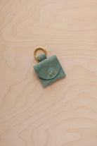 A small eucalyptus coloured sage green suede pouch bag charm with a gold coloured clip ring fastening, displayed on a light wood grain background.
