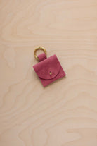 A small faded rose pink coloured suede pouch bag charm with a gold coloured clip ring fastening, displayed on a light wood grain background.