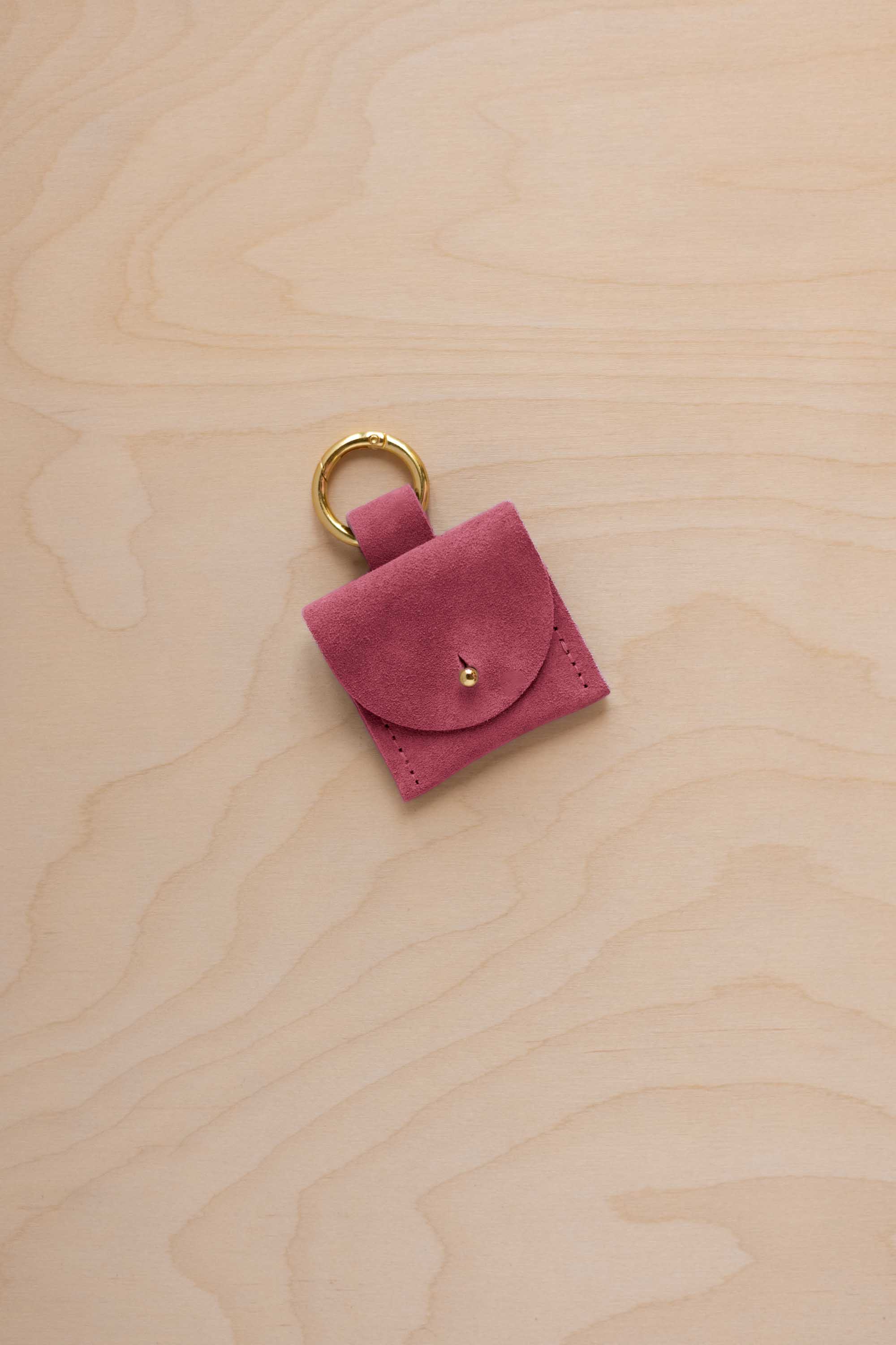 A small faded rose pink coloured suede pouch bag charm with a gold coloured clip ring fastening, displayed on a light wood grain background.
