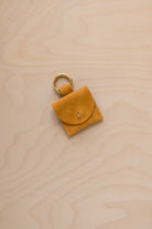 A small golden ochre mustard yellow yellow suede pouch bag charm with a gold coloured clip ring fastening, displayed on a light wood grain background.