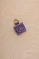 A small lavender coloured suede pouch bag charm with a gold coloured clip ring fastening, displayed on a light wood grain background.