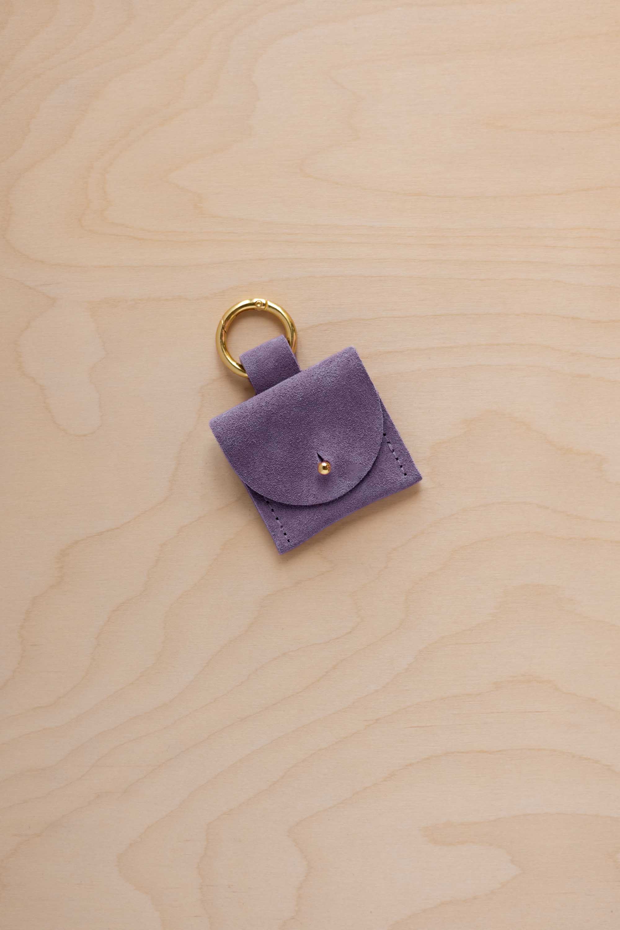 A small lavender coloured suede pouch bag charm with a gold coloured clip ring fastening, displayed on a light wood grain background.