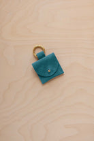 A small marine teal coloured suede pouch bag charm with a gold coloured clip ring fastening, displayed on a light wood grain background.