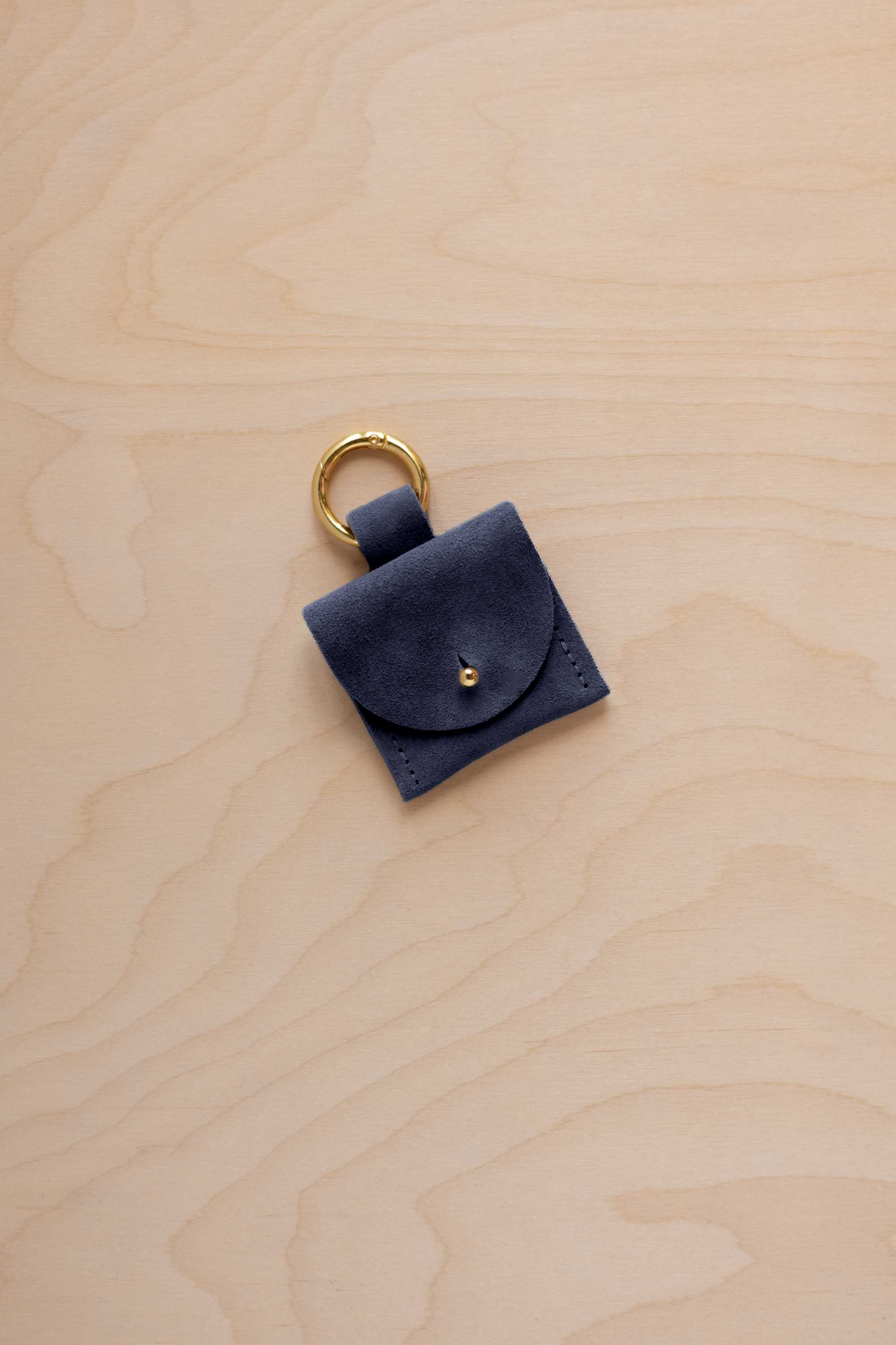 A small midnight navy blue coloured suede pouch bag charm with a gold coloured clip ring fastening, displayed on a light wood grain background.