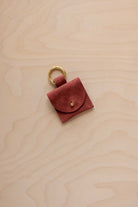 A small rust red coloured suede pouch bag charm with a gold coloured clip ring fastening, displayed on a light wood grain background.