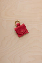 A small scarlet red coloured suede pouch bag charm with a gold coloured clip ring fastening, displayed on a light wood grain background.