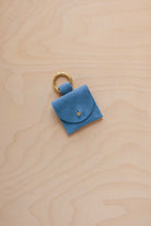 A small sky blue coloured suede pouch bag charm with a gold coloured clip ring fastening, displayed on a light wood grain background.