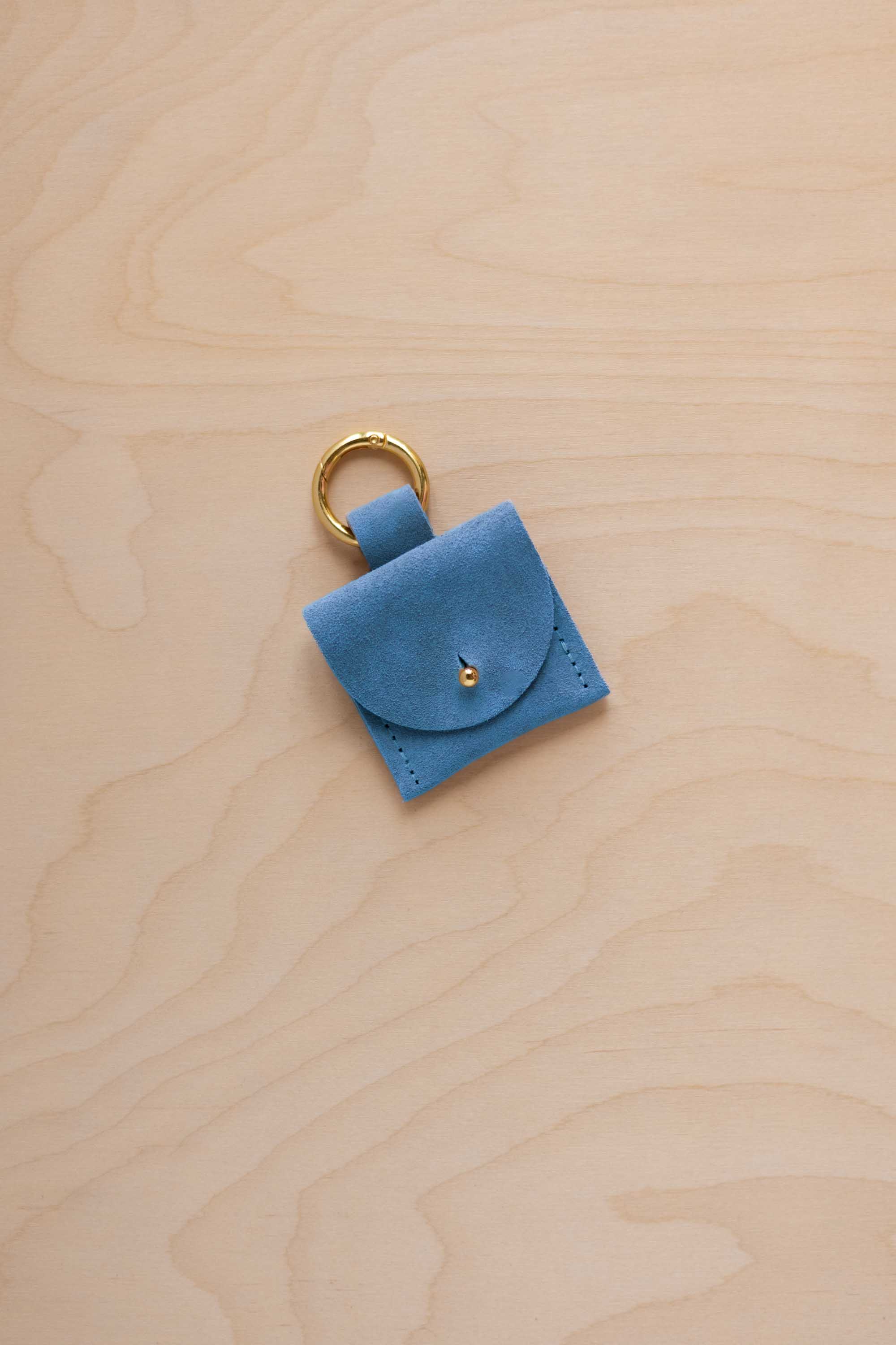 A small sky blue coloured suede pouch bag charm with a gold coloured clip ring fastening, displayed on a light wood grain background.