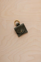 A small soft olive green coloured suede pouch bag charm with a gold coloured clip ring fastening, displayed on a light wood grain background.