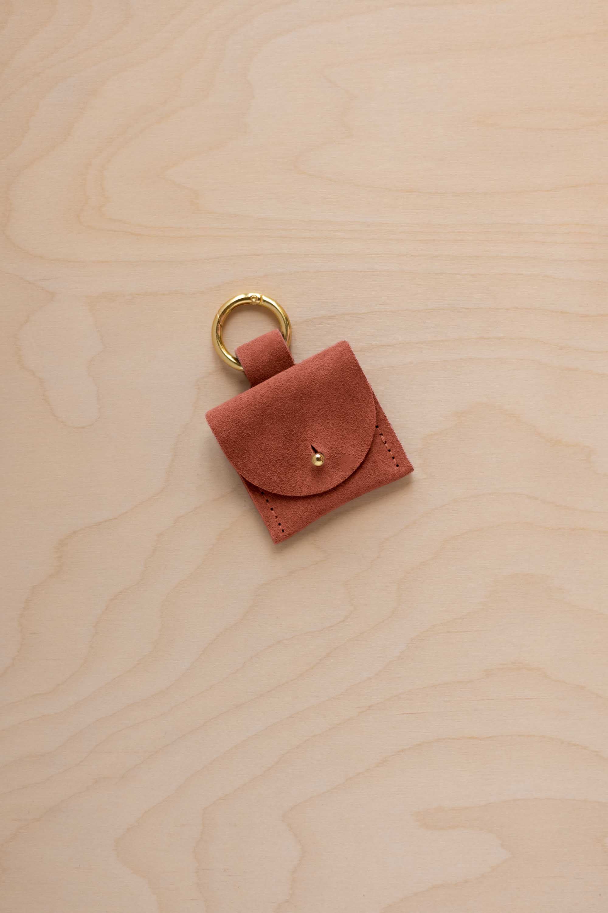 A small terracotta brown coloured suede pouch bag charm with a gold coloured clip ring fastening, displayed on a light wood grain background.