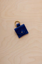 A small ultramarine blue coloured suede pouch bag charm with a gold coloured clip ring fastening, displayed on a light wood grain background.