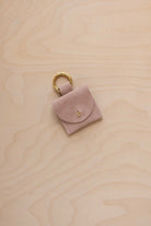 A small beige suede pouch bag charm with a gold coloured clip ring fastening, displayed on a light wood grain background.