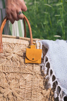 A close-up of a woven straw tote bag with a small mustard yellow suede pouch with gold clip hanging from a tan leather handle, held by a hand outdoors with a white fringed blanket draped over the side.