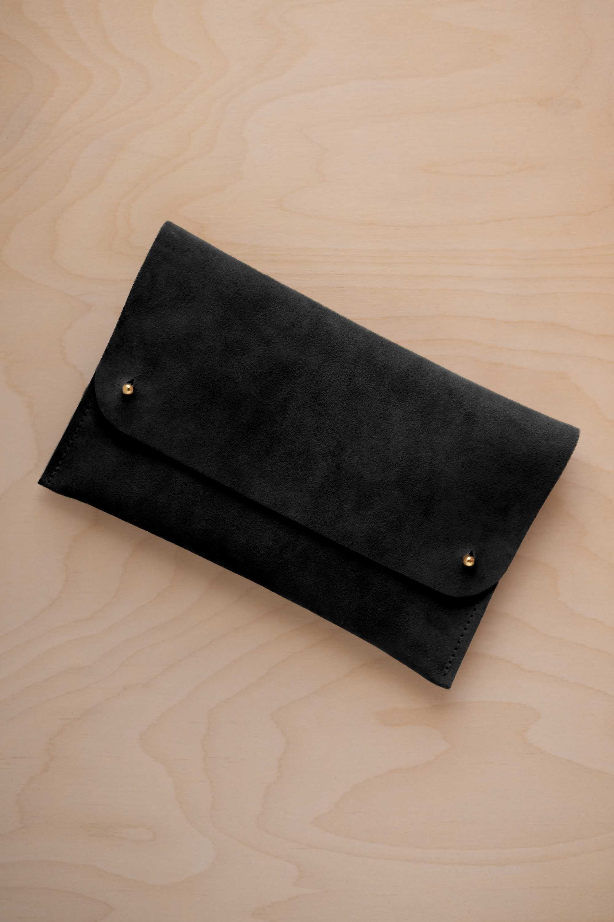 A black suede clutch with small gold coloured metal studs, displayed diagonally on a light wood grain surface.