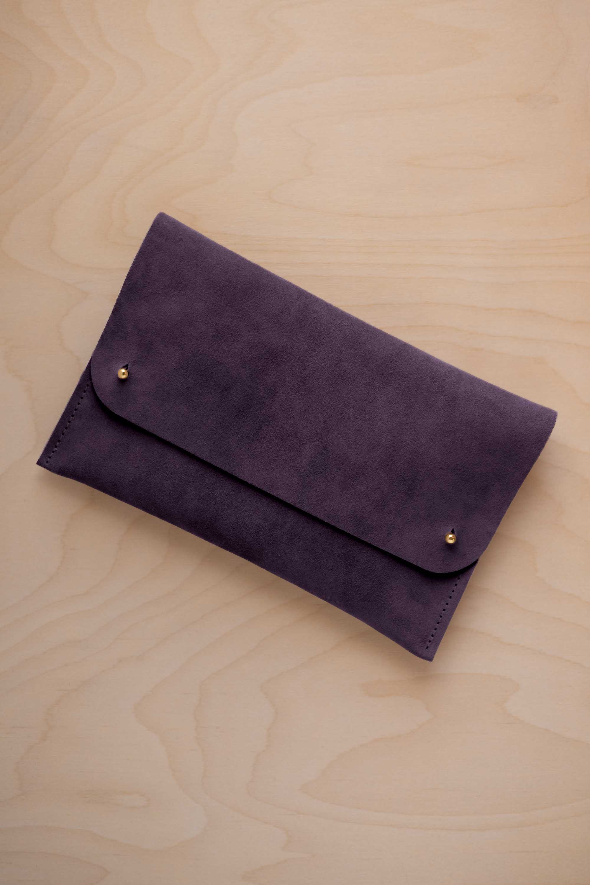 A blackcurrant dark purple suede clutch with small gold coloured metal studs, displayed diagonally on a light wood grain surface.