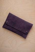 A blackcurrant dark purple suede clutch with small gold coloured metal studs, displayed diagonally on a light wood grain surface.