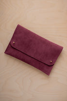 A burgundy suede clutch with small gold coloured metal studs, displayed diagonally on a light wood grain surface.