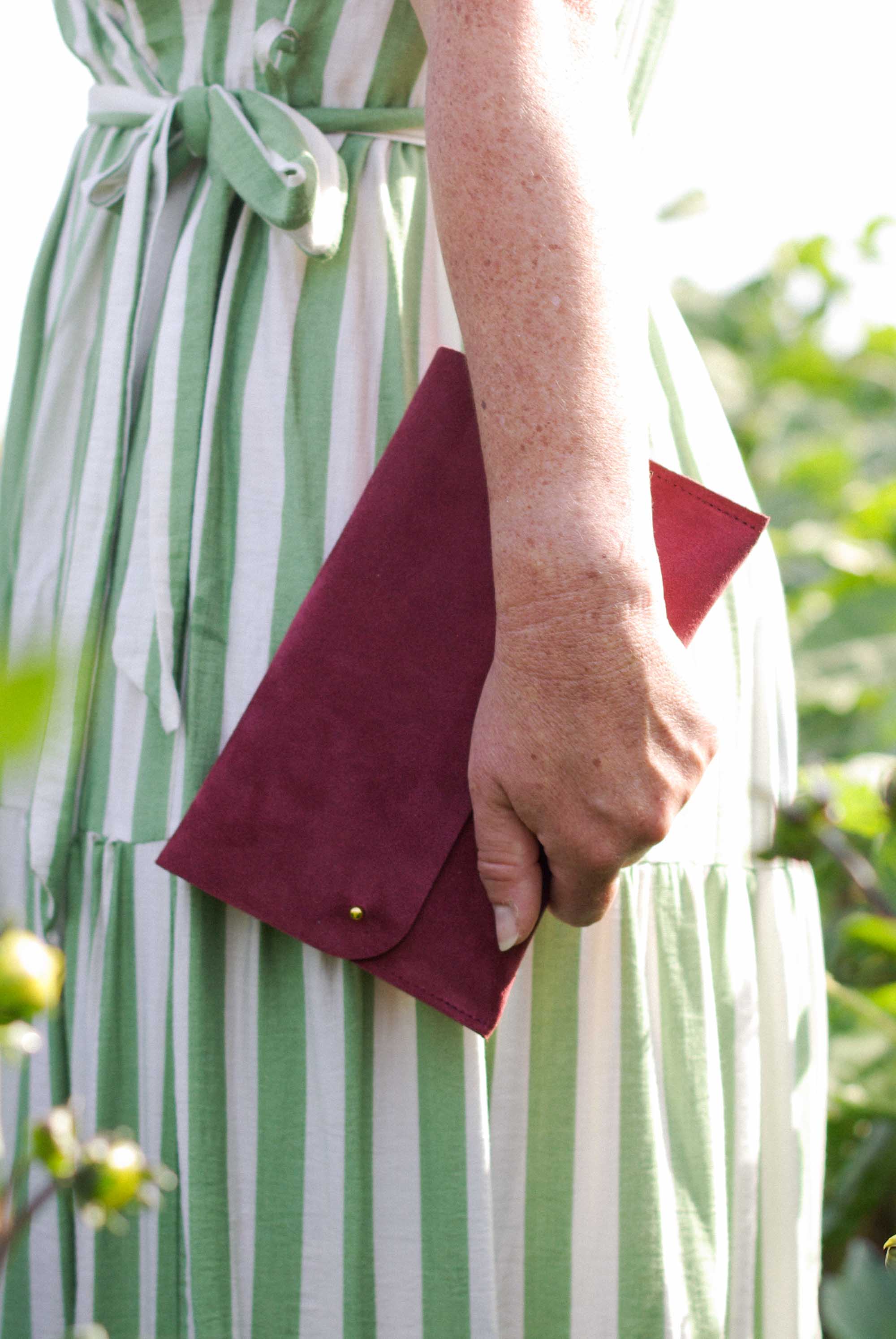 A close-up of a person wearing a green and white striped dress, holding a burgundy suede envelope clutch with a gold snap button.