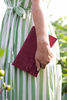 A close-up of a person wearing a green and white striped dress, holding a burgundy suede envelope clutch with a gold snap button.
