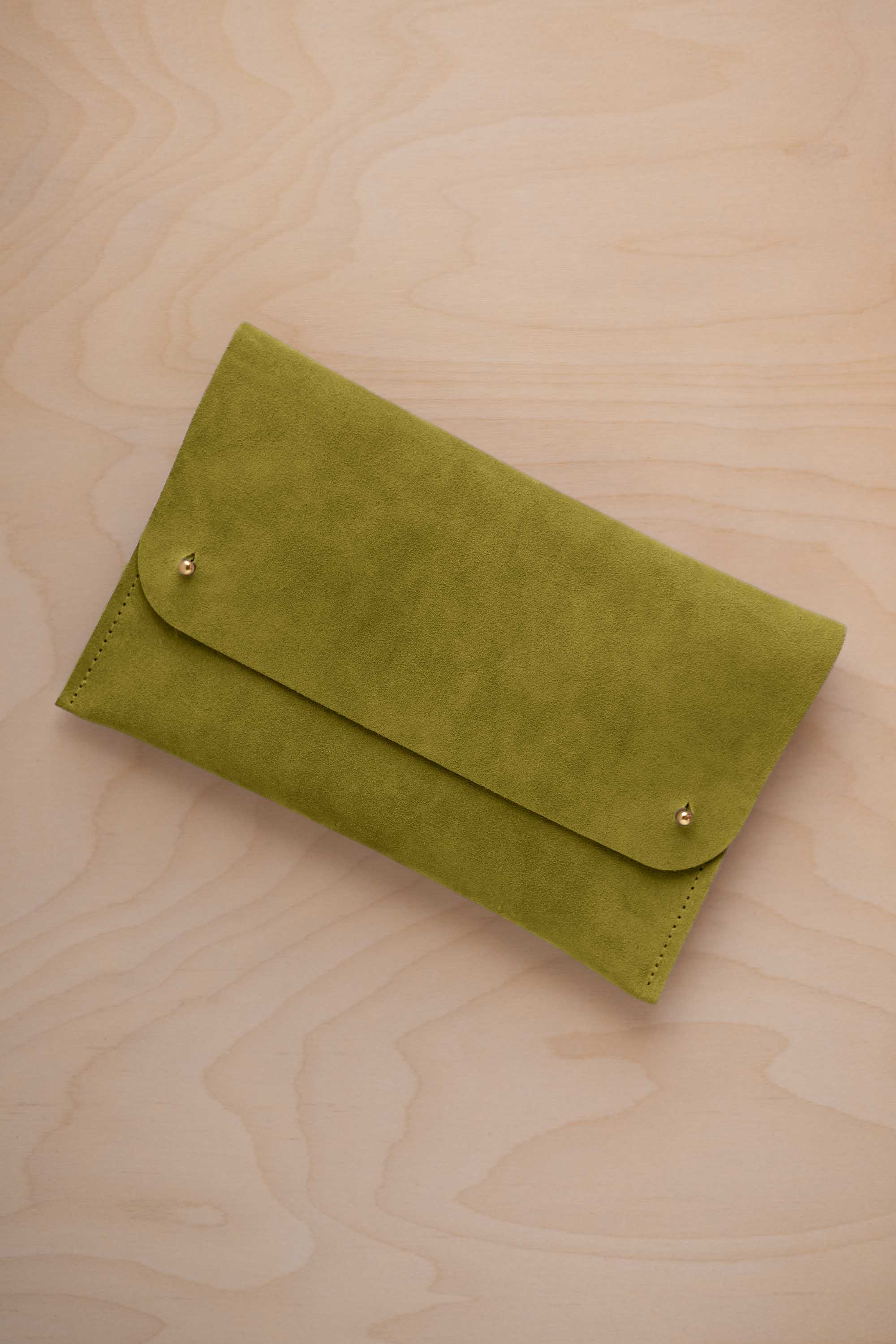A chartreuse lime green coloured suede clutch with small gold coloured metal studs, displayed diagonally on a light wood grain surface.