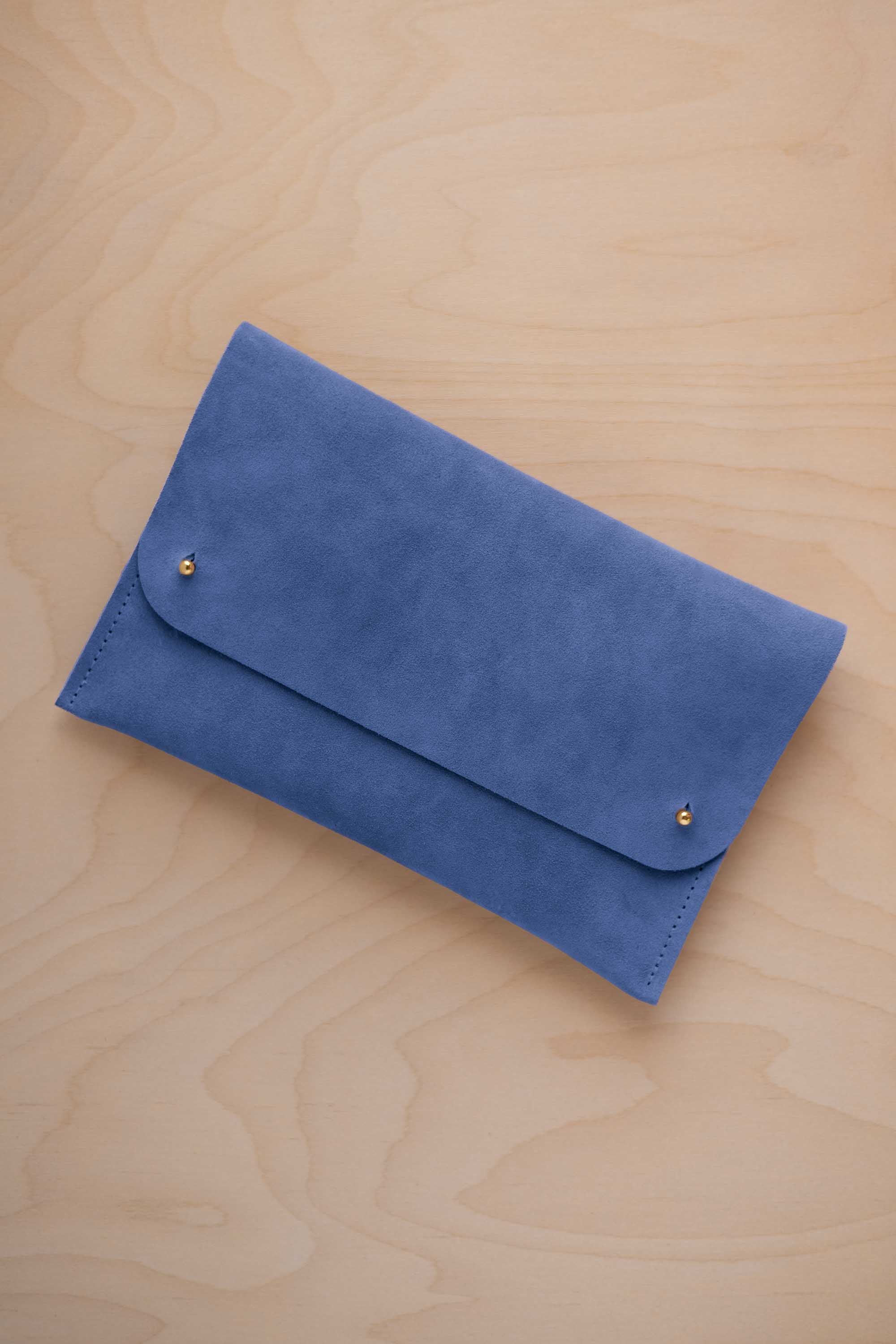 A cobalt blue suede clutch with small gold coloured metal studs, displayed diagonally on a light wood grain surface.