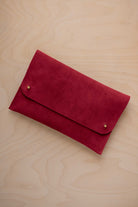 A crimson red suede clutch with small gold coloured metal studs, displayed diagonally on a light wood grain surface.