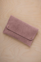 A dusty mauve grey suede clutch with small gold coloured metal studs, displayed diagonally on a light wood grain surface.
