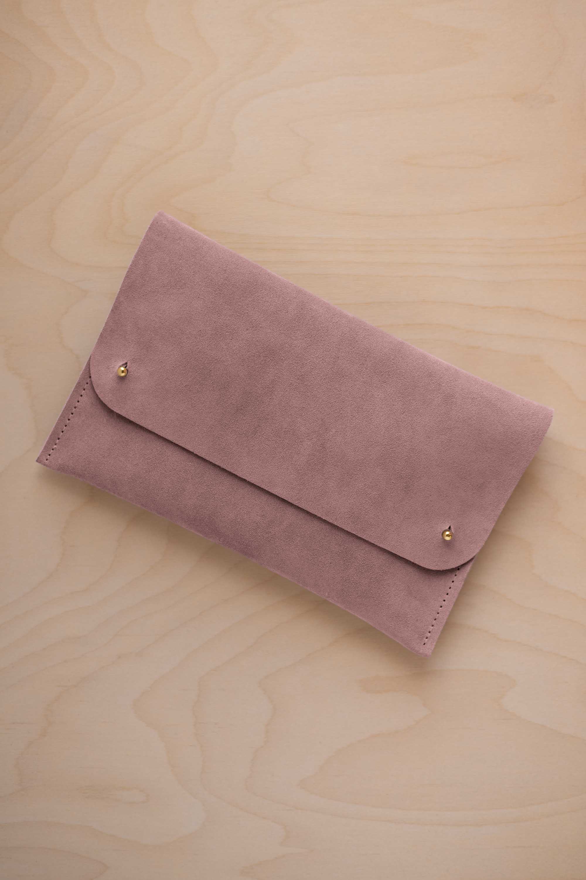A dusty mauve grey suede clutch with small gold coloured metal studs, displayed diagonally on a light wood grain surface.
