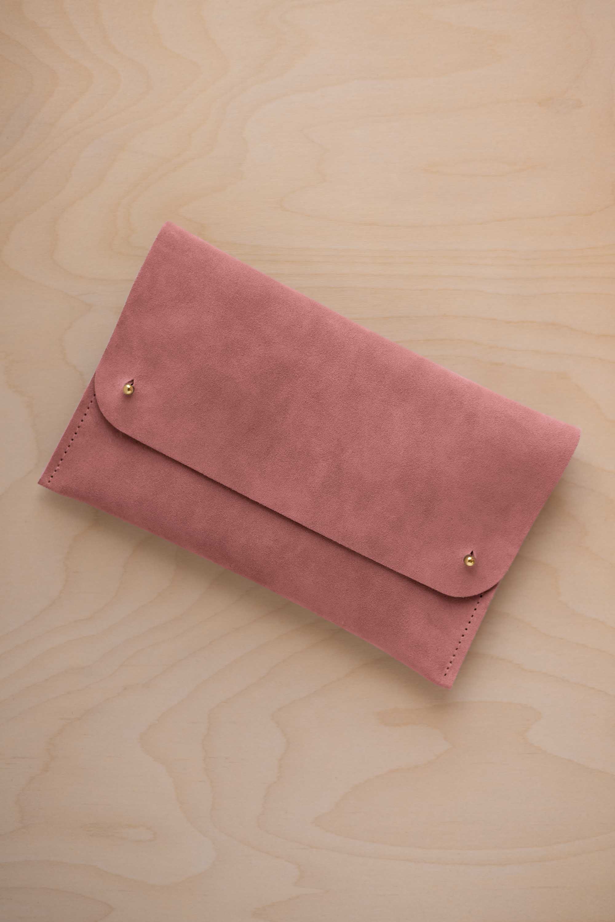 A dusty rose pink coloured suede clutch with small gold coloured metal studs, displayed diagonally on a light wood grain surface.