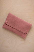A dusty rose pink coloured suede clutch with small gold coloured metal studs, displayed diagonally on a light wood grain surface.