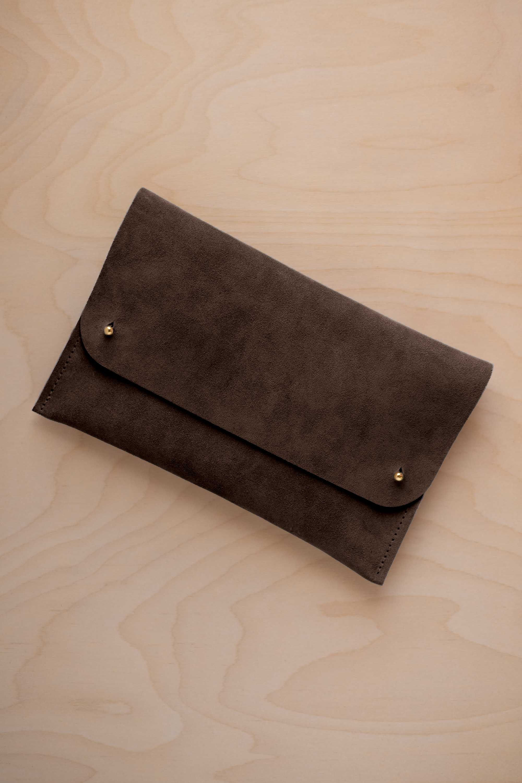 An espresso brown suede clutch with small gold coloured metal studs, displayed diagonally on a light wood grain surface.