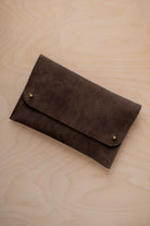 An espresso brown suede clutch with small gold coloured metal studs, displayed diagonally on a light wood grain surface.