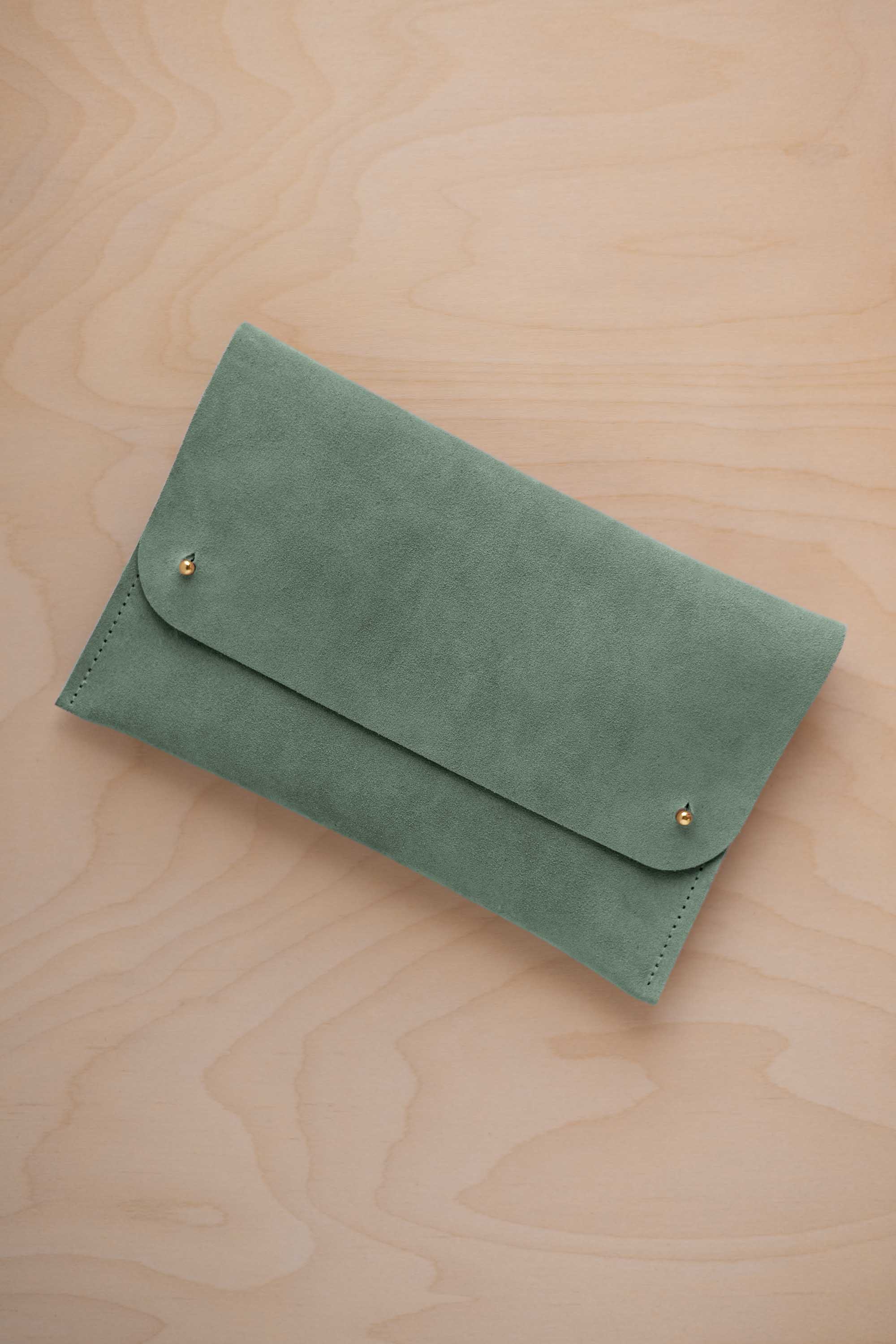 A eucalyptus sage green coloured suede clutch with small gold coloured metal studs, displayed diagonally on a light wood grain surface.