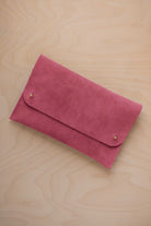 A faded rose pink suede clutch with small gold coloured metal studs, displayed diagonally on a light wood grain surface.