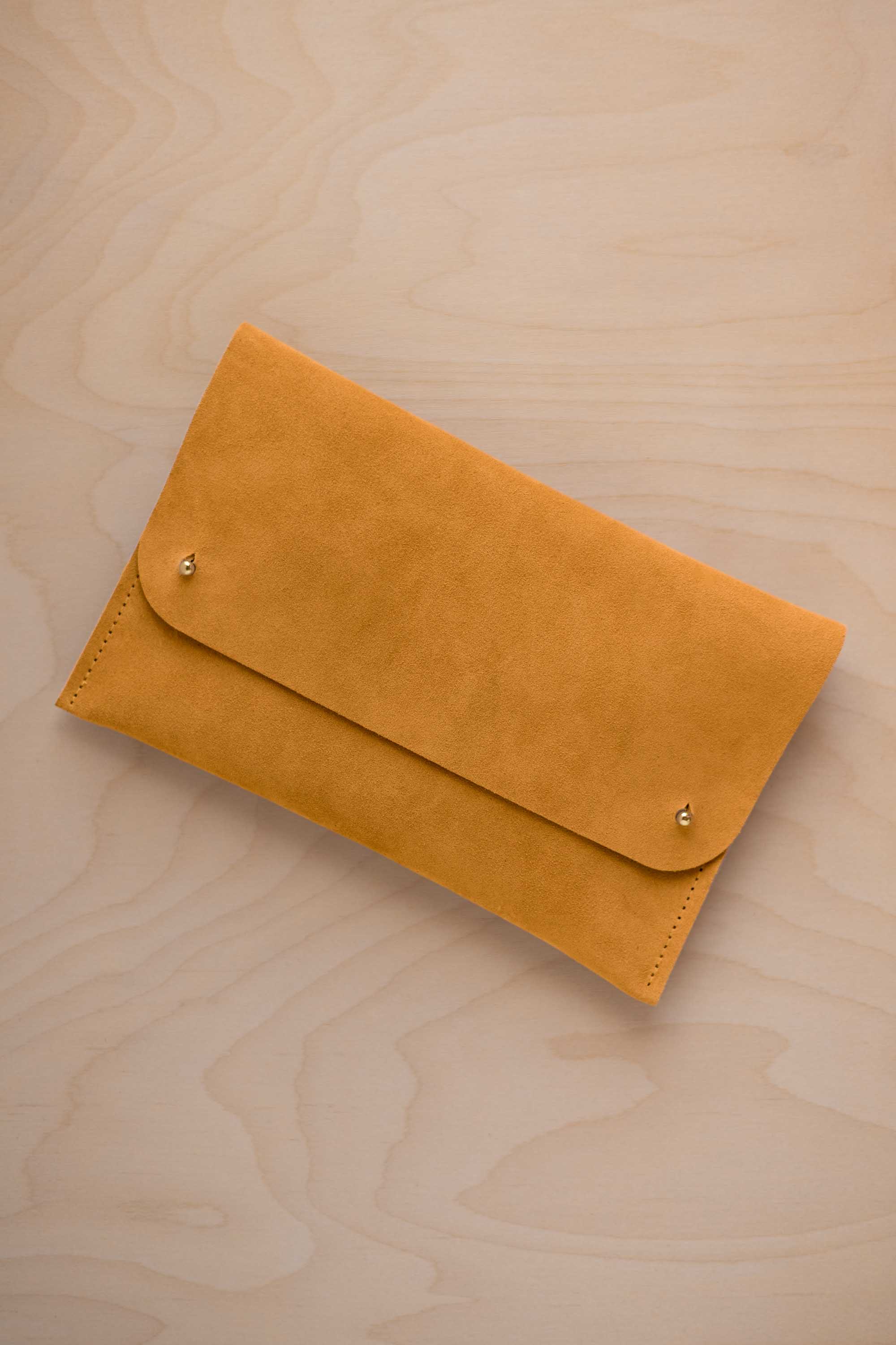 A golden ochre mustard yellow coloured suede clutch with small gold coloured metal studs, displayed diagonally on a light wood grain surface.