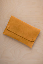 A golden ochre mustard yellow coloured suede clutch with small gold coloured metal studs, displayed diagonally on a light wood grain surface.