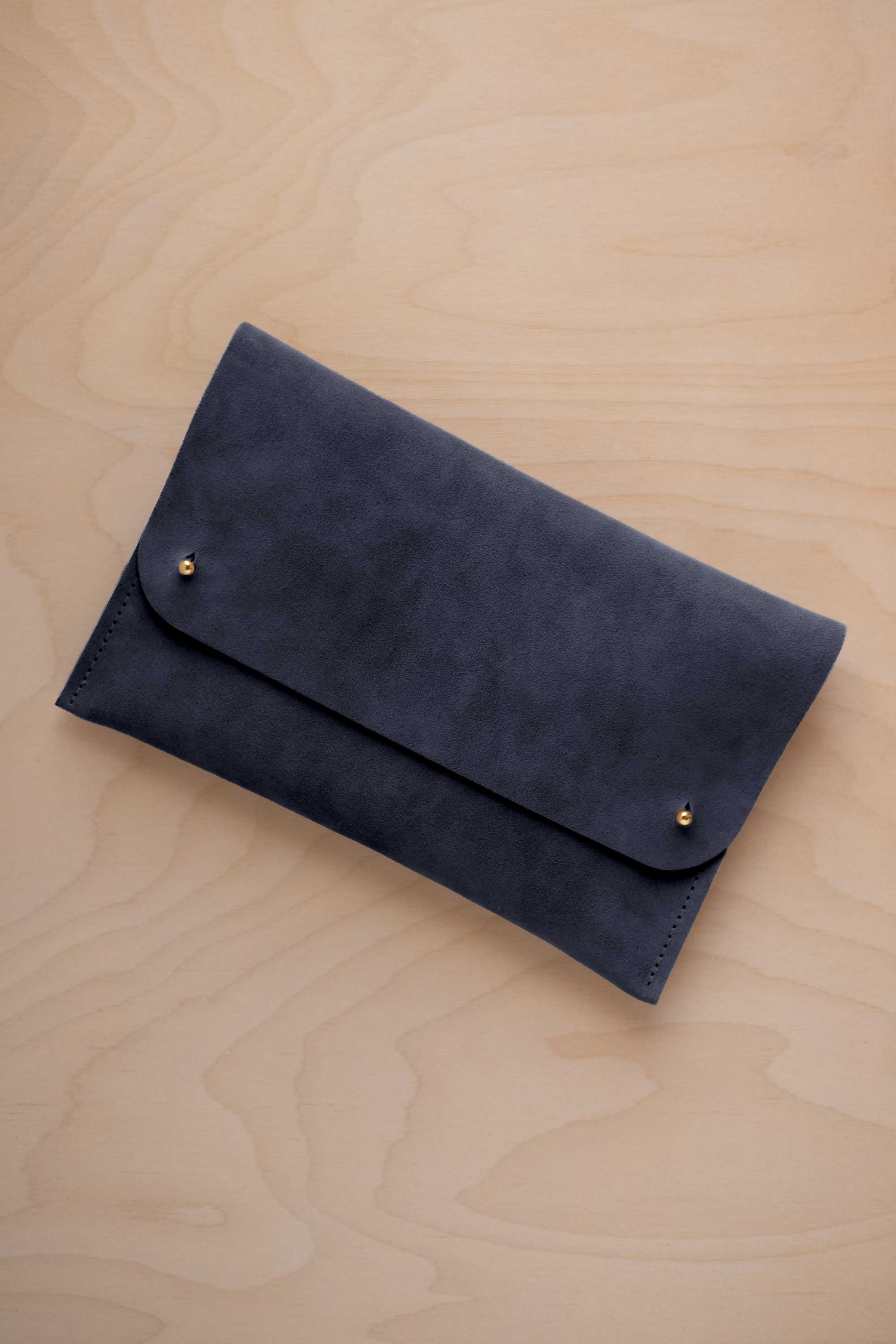 A midnight blue coloured suede clutch with small gold coloured metal studs, displayed diagonally on a light wood grain surface.