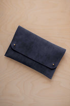 A midnight blue coloured suede clutch with small gold coloured metal studs, displayed diagonally on a light wood grain surface.