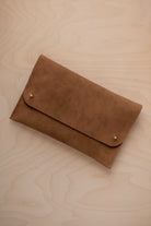 A mocha brown coloured suede clutch with small gold coloured metal studs, displayed diagonally on a light wood grain surface.