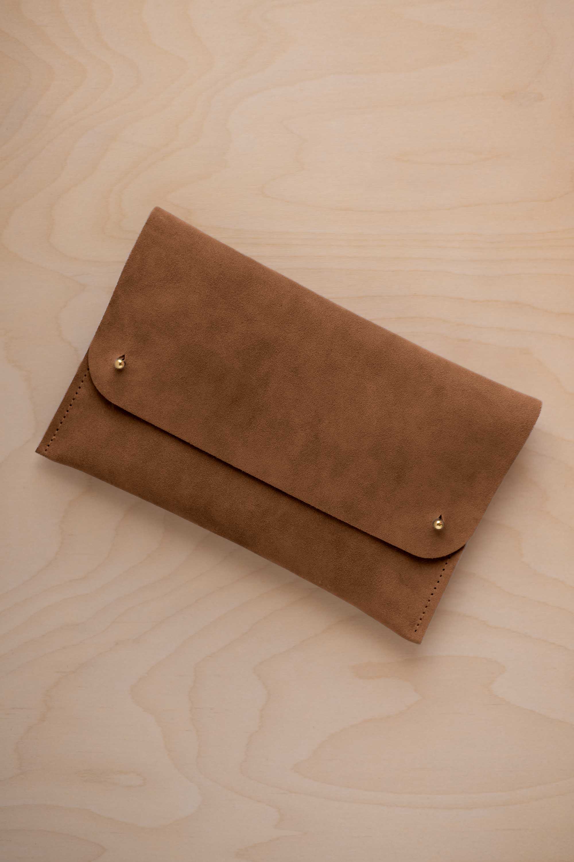 A mocha brown coloured suede clutch with small gold coloured metal studs, displayed diagonally on a light wood grain surface.