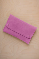 A peony pink suede clutch with small gold coloured metal studs, displayed diagonally on a light wood grain surface.