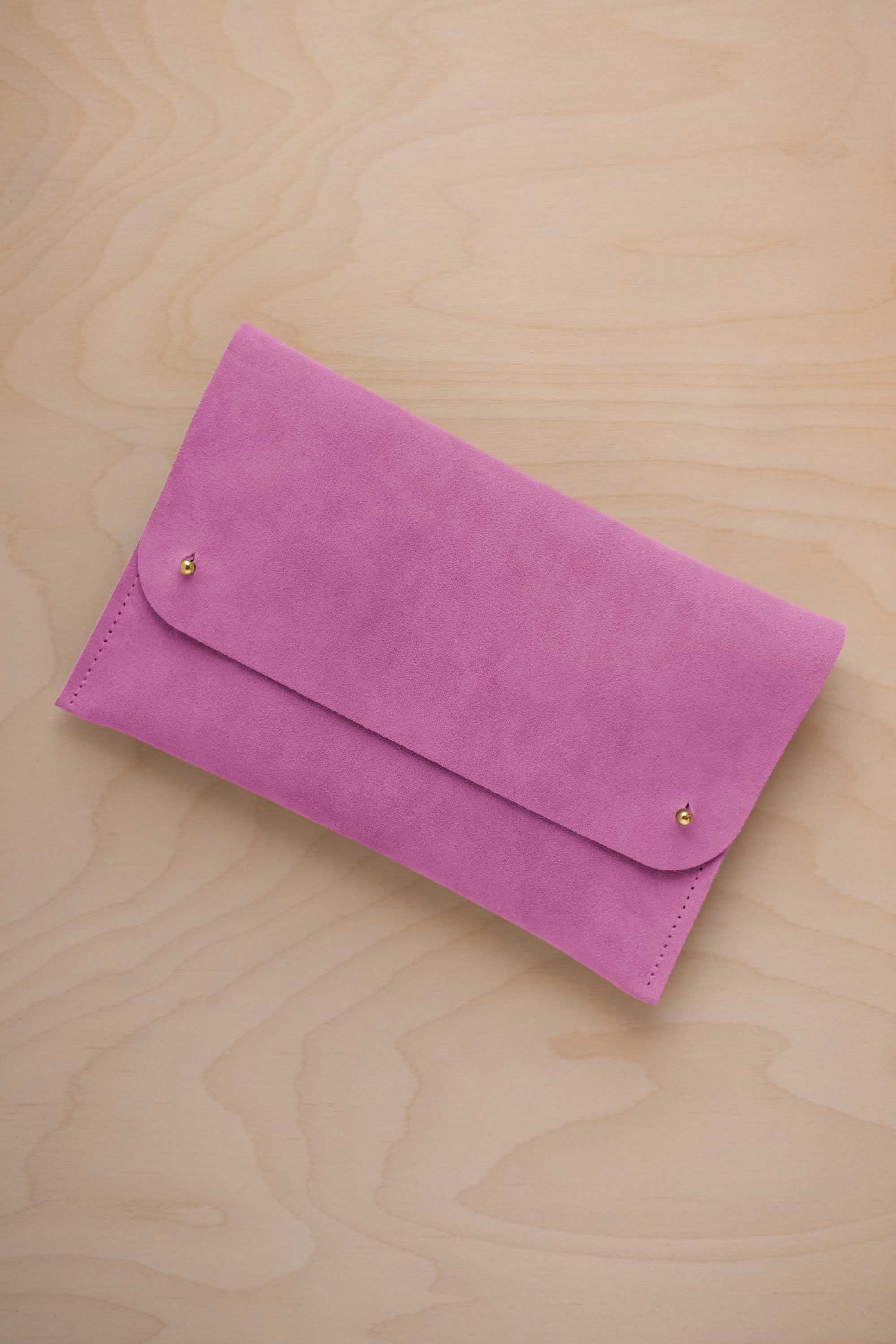 A peony pink suede clutch with small gold coloured metal studs, displayed diagonally on a light wood grain surface.