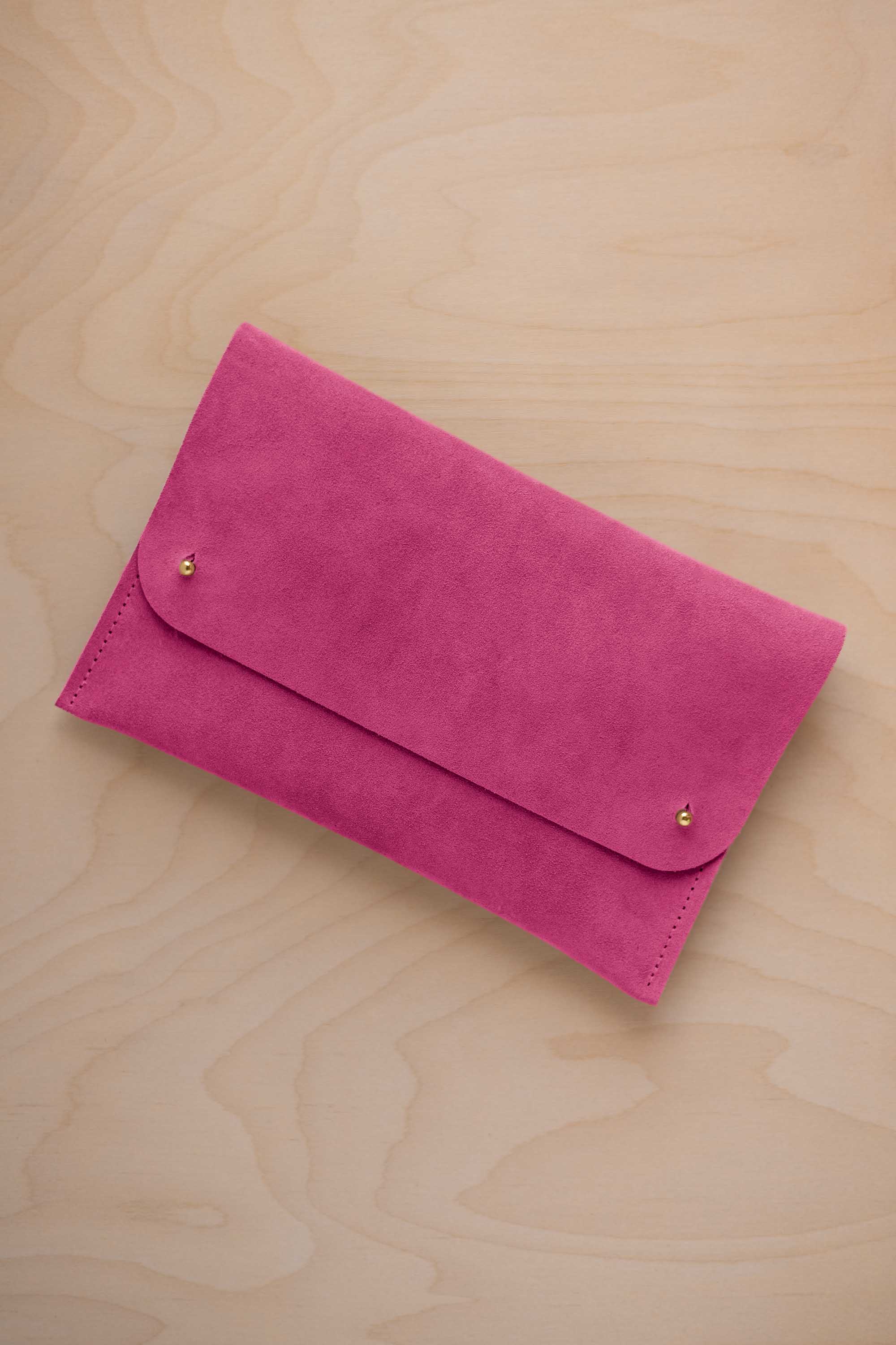 A raspberry pink coloured suede clutch with small gold coloured metal studs, displayed diagonally on a light wood grain surface.