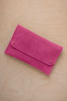 A raspberry pink coloured suede clutch with small gold coloured metal studs, displayed diagonally on a light wood grain surface.