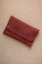 A rust red suede clutch with small gold coloured metal studs, displayed diagonally on a light wood grain surface.