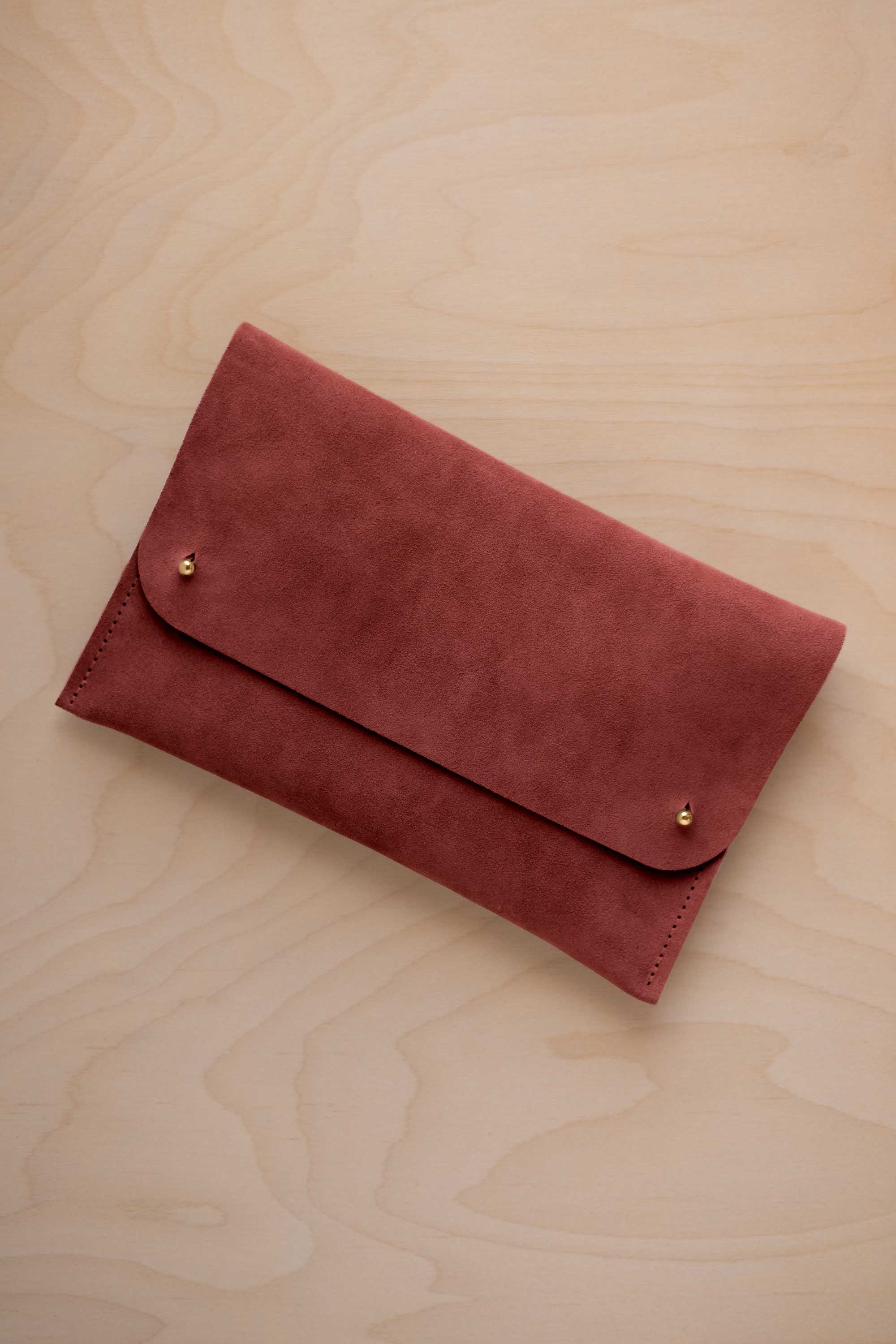 A rust red suede clutch with small gold coloured metal studs, displayed diagonally on a light wood grain surface.