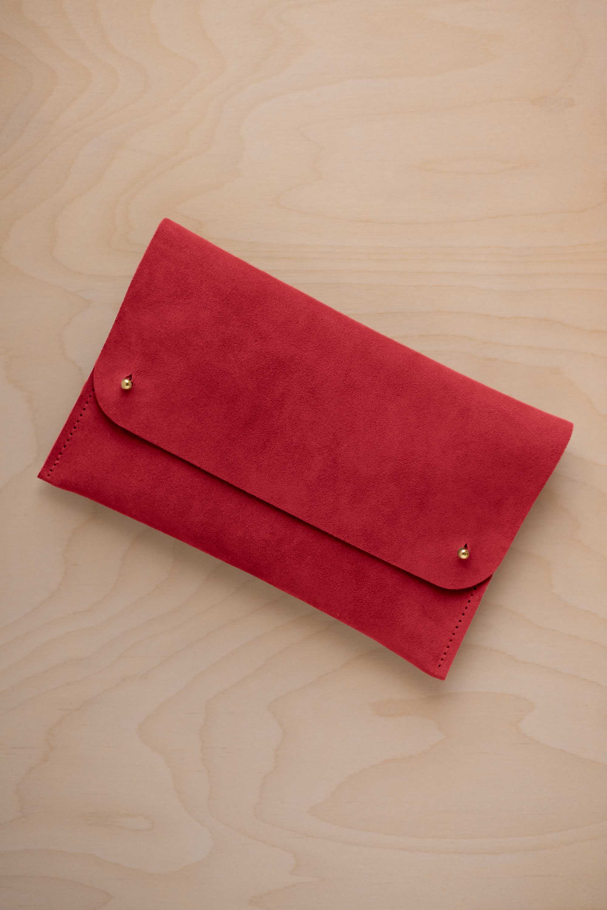 A scarlet red suede clutch with small gold coloured metal studs, displayed diagonally on a light wood grain surface.