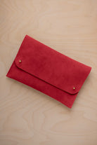 A scarlet red suede clutch with small gold coloured metal studs, displayed diagonally on a light wood grain surface.