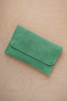 A seafoam sage green coloured suede clutch with small gold coloured metal studs, displayed diagonally on a light wood grain surface.