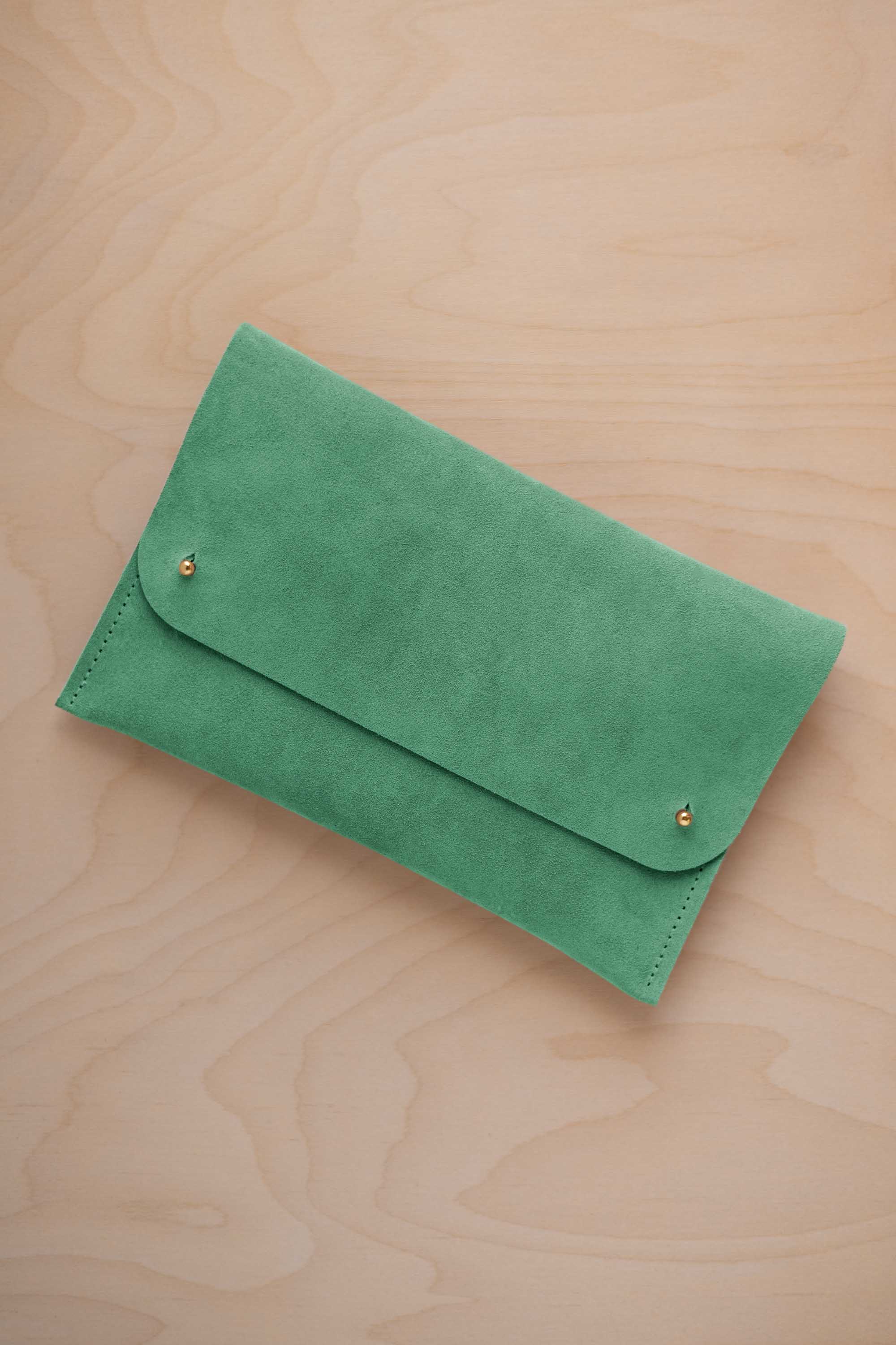 A seafoam sage green coloured suede clutch with small gold coloured metal studs, displayed diagonally on a light wood grain surface.
